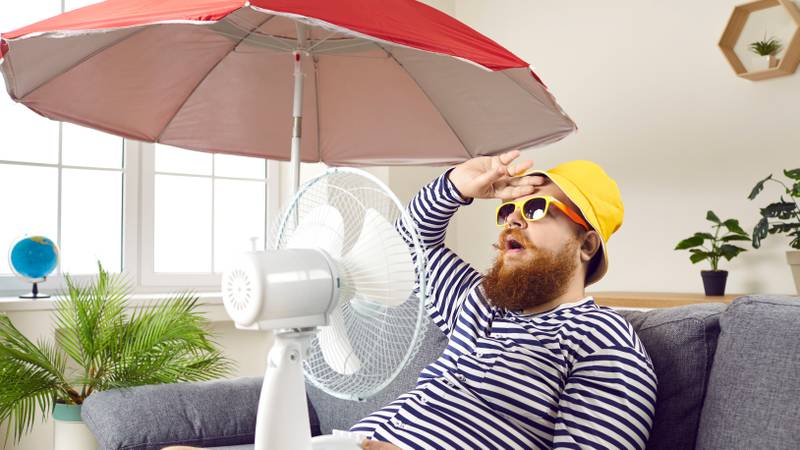 Funny sweaty bearded man in swimsuit sitting at home, suffering from crazy summer heat, wiping sweat off forehead, holding electric fan, wishing for heatwave to stop and fresh breeze to blow