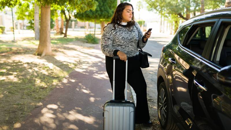 A woman waiting for a ride holding a cellphone and the handle of a suitcase.